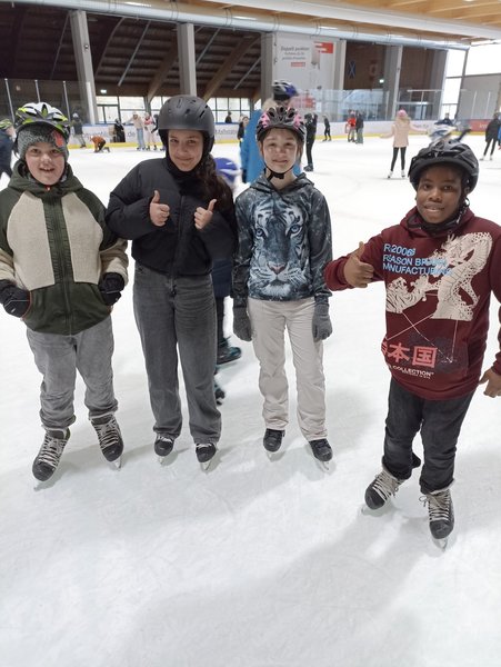 Vier Kinder mit Helm stehen lachend in Schlittschuhen auf dem Eis. Ein Mädchen reckt die Daumen nach oben.