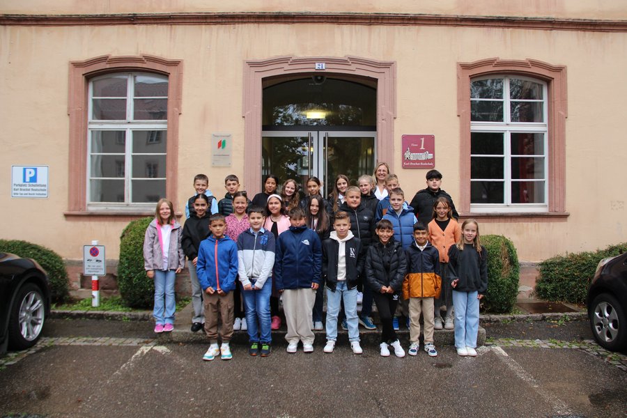 Die Klasse 5e mit ihrer Klassenlehrerin Frau Mayer Die Schulkinder stehen in mehreren Reihen auf Treppenstufen vor dem Schuleingang und lächeln in die Kamera.