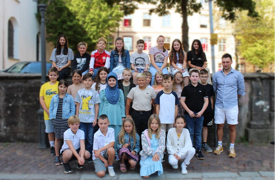 Schülerinnen und Schüler stehen für ein Klassenfoto zusammen. Die vordere Reihe kniet, dahinter steht eine Reihe die letzte Reihe sitzt auf einer Mauer. Im Hintergrund sieht man die Schule.
