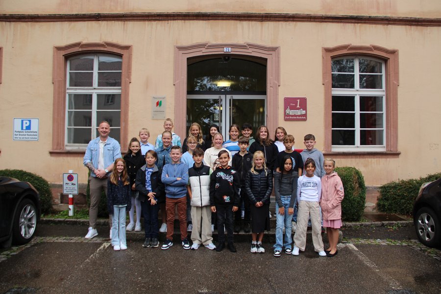 Die Klasse 5d mit ihrem Klassenlehrer Herr Scholl Die Schulkinder stehen in mehreren Reihen auf Treppenstufen vor dem Schuleingang und lächeln in die Kamera.