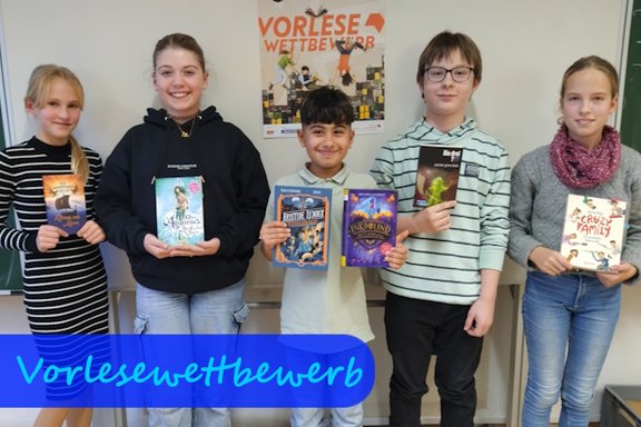 Vor einer weißen Wand mit einem Plakat "Vorlesewettbewerb" stehen drei Mädchen und zwei Jungs nebeneinander und halten jeweils ein Buch Richtung Kamera.