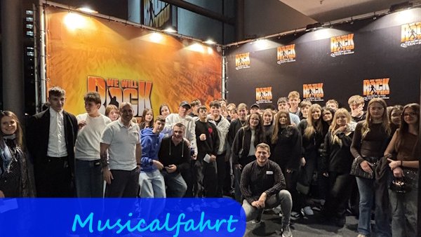 Vor einem großen Plakat des Musicals "We will rock you" stehen Jugendliche zu einem Gruppenbild zusammen.