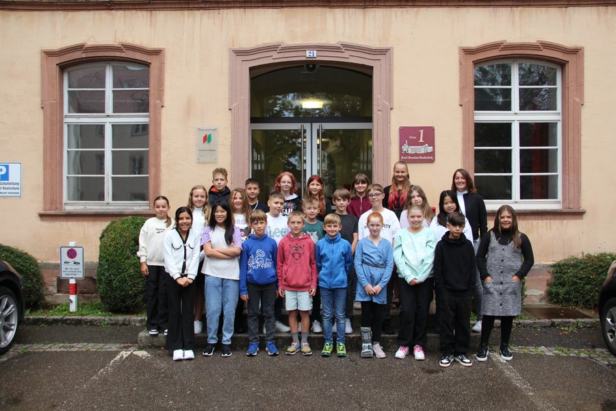 Die Klasse 5a mit ihrer Klassenlehrerin Frau Spadinger Die Schulkinder stehen in mehreren Reihen auf Treppenstufen vor dem Schuleingang und lächeln in die Kamera.