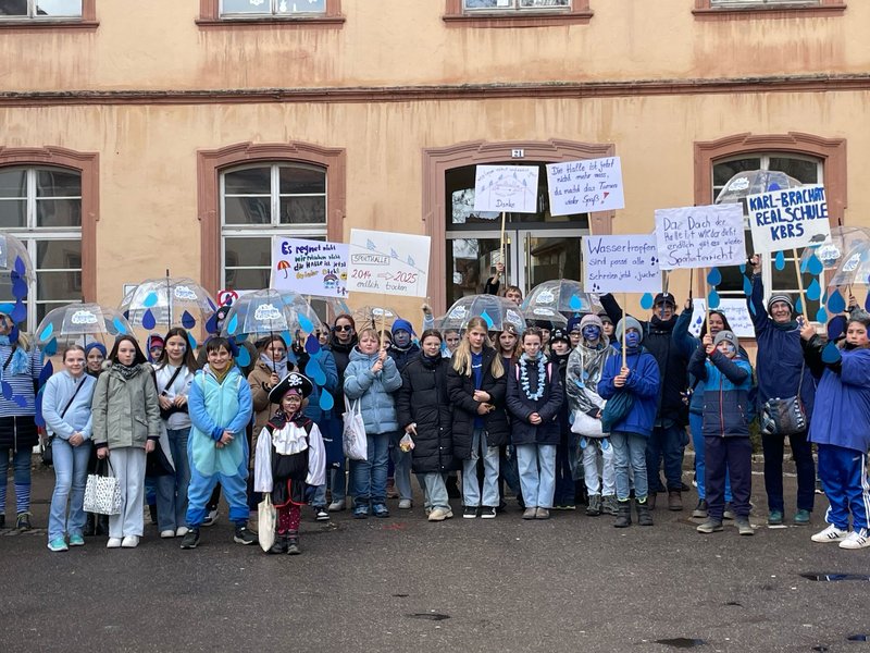 Gruppenbild vor dem Eingang einer Schule. Die Kinder und Erwachsenen halten Plakate und Schirme in die Höhe. Die meisten sind blau gekleidet.