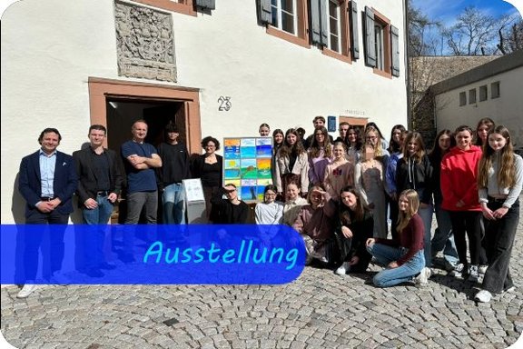 Erwachsene und Jugendliche stehen vor dem Eingang eines alten, restaurierten Hauses. In der Mitte ist ein Aufsteller mit verschiedenen Gemälden der Jugendlichen.