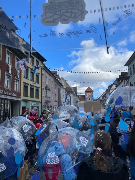 Von hinten sieht man viele Kinder unter transparenten Regenschirmen in Richtung eines Stadttores laufen.