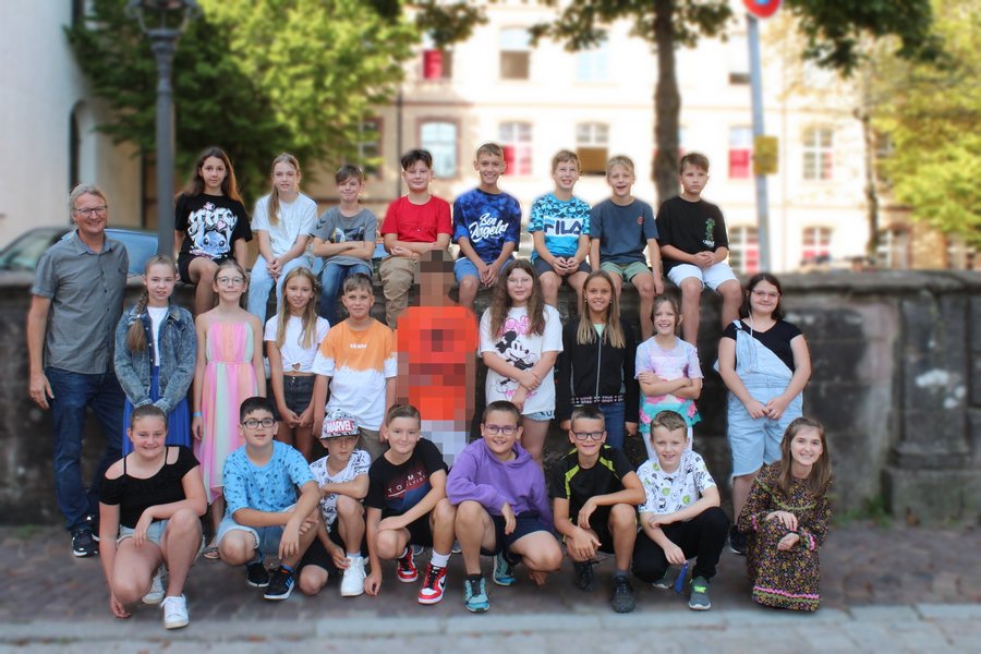 Schülerinnen und Schüler stehen für ein Klassenfoto zusammen. Die vordere Reihe kniet, dahinter steht eine Reihe die letzte Reihe sitzt auf einer Mauer. Im Hintergrund sieht man die Schule.
