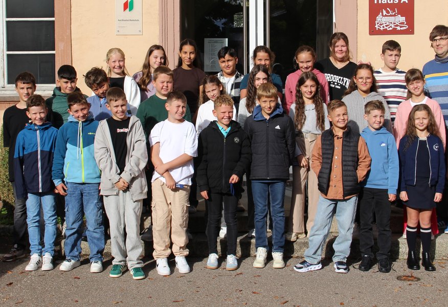 Vor dem Eingang der Schule hat sich eine fünfte Klasse mit der Klassenlehrkraft zum Gruppenfoto aufgestellt.