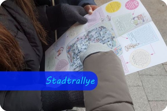 In der Bildmitte sieht man einen aufgeschlagenen Stadtplan, der von einem Kind in der Hand gehalten wird. Eine weitere Kinderhand zeigt auf einen Punkt auf der Karte.