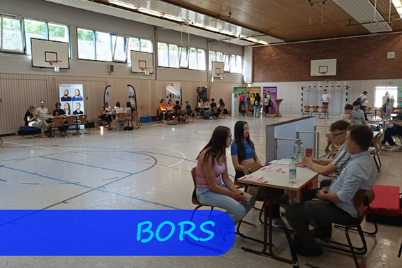 In einer Turnhalle stehen reihum am Rand Tische an denen Schülerinnen und Schüler mit Vertretern unterschiedlicher Berufe im Gespräch sind.