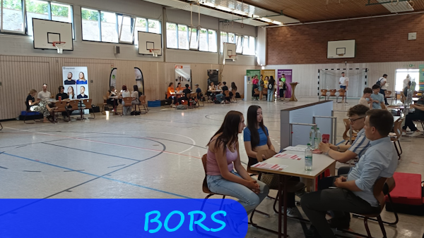 In einer Turnhalle stehen reihum am Rand Tische an denen Schülerinnen und Schüler mit Vertretern unterschiedlicher Berufe im Gespräch sind.