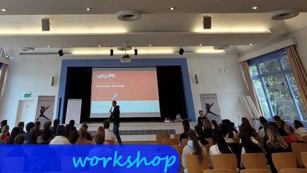 Im Hintergrund sieht man eine große Leinwand, auf der eine Präsentation läuft. Davor ist ein Mann mit einem Mikrophon. Von hinten sieht man die Zuhörer sitzen.
