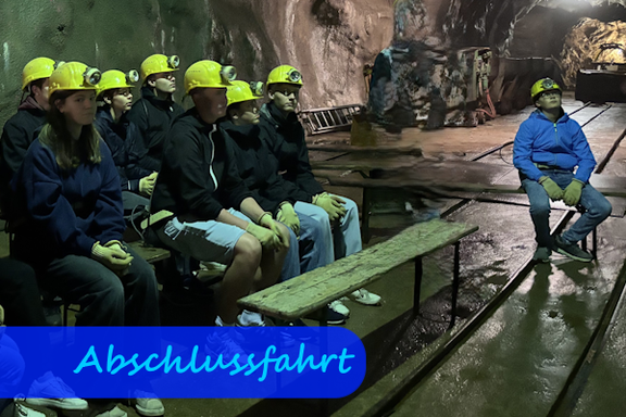 In einem Stollen sitzen Schülerinnen und Schüler mit gelben Helmen auf Bänken und schauen nach rechts an der Kamera vorbei in Richtung eines Vortrags.