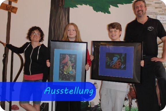 Vier Personen stehen nebeneinander. In der Mitte stehen ein Mädchen und ein Junge, die jeweils ein gerahmtes Bild in Richtung Kamera halten.