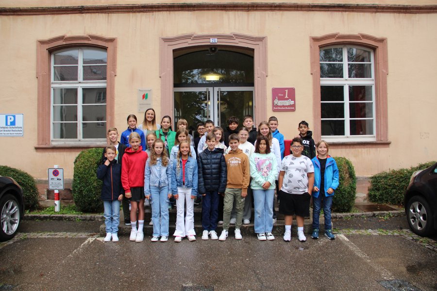 Die Klasse 5b mit ihrer Klassenlehrerin Frau Walter Die Schulkinder stehen in mehreren Reihen auf Treppenstufen vor dem Schuleingang und lächeln in die Kamera.