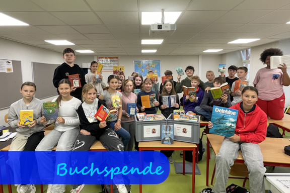 Auf Tischen sitzen Jungen und Mädchen mit Büchern in der Hand. In der Bildmitte stehen zwei graue Plastikkisten mit Büchern darin.