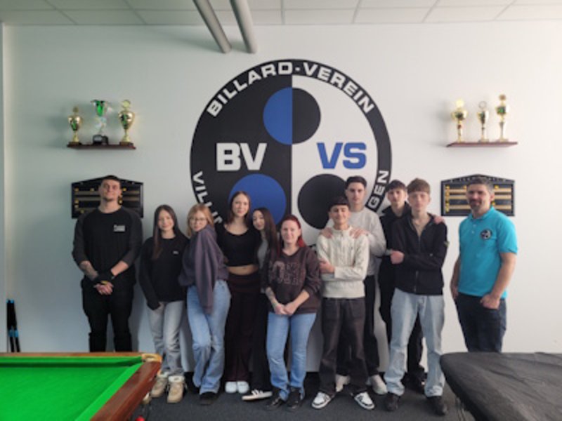 Vor einer weißen Wand mit einem großen Logo des Bllardvereins haben sich Jungen und Mädchen zu einem Grupenfoto aufgestellt. Links und rechts von ihnen stehen die beiden Trainer.