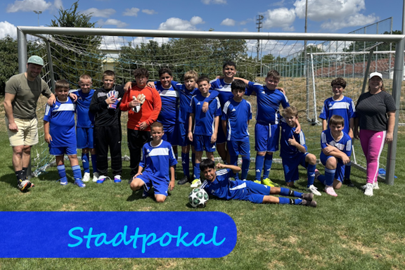 Vor einem Fußballtor haben sich Jungen in blauen Trikots zu einem Mannschaftsfoto aufgestellt, davor kniet und liegt jeweils ein Jungen. Links und rechts stehen eine männliche und eine weibliche Lehrkraft.