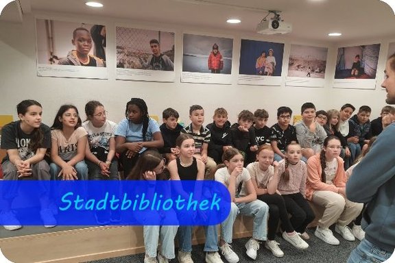 Am rechten Rand sieht man das Profil eines Mannes. Er spricht zu Jungen und Mädchen, die auch Potesten sitzen und ihm zuhören.