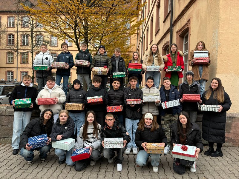 In drei Reihen stehen Schülerinnen und Schüler der Klassenstufe 7 vor und auf einer kleinen Mauer und schauen in die Kamera. In den Händen halten sie weihnachtlich verpackte Päckchen. Rechts daneben steht ihre Lehrerin, ebenfalls mit Päckchen in der Hand.