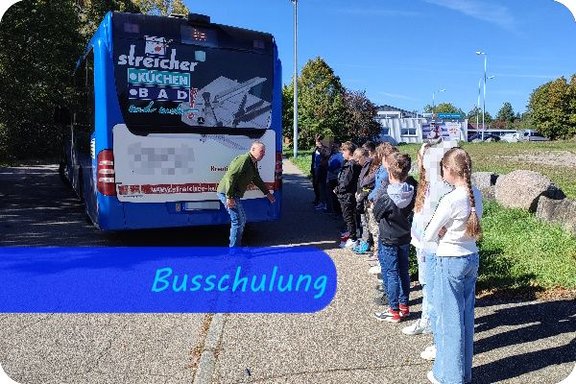 Das Heck eines Busses steht über den Fußweg hinaus. Dahinter beugt sich ein Mann, um den Überhang zu zeigen. Eine Reihe von Kindern steht in einer Linie mit Abstand daneben und hört zu.