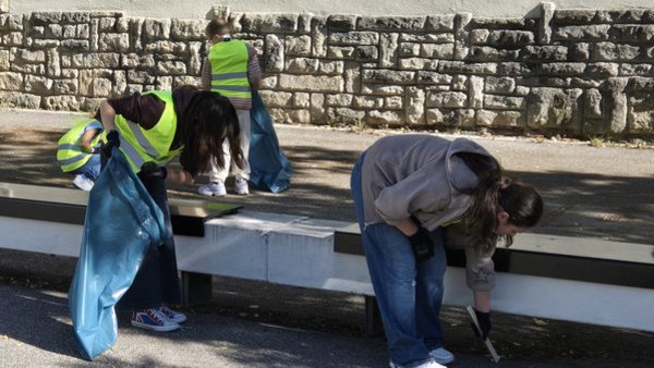 Vier Kinder sammeln Müll auf. Sie haben einen Müllsack und Zangen. Drei tragen gelbe Warnwesten. Im Vordergrund beugen sich zwei vor einer Sitzbank nach Müll.  
