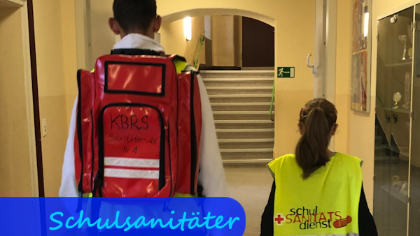 In einem Schulflur gehen ein größerer Junge und ein kleineres Mädchen vom Fotografen weg. Das Mädchen trägt eine gelbe Warnweste mit der Aufschrift "Schulsanitätsdienst". Der Junge träg einen roten Erste-Hilfe-Rucksack auf dem Rücken.