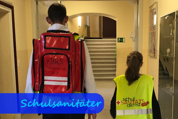 In einem Schulflur gehen ein größerer Junge und ein kleineres Mädchen vom Fotografen weg. Das Mädchen trägt eine gelbe Warnweste mit der Aufschrift "Schulsanitätsdienst". Der Junge träg einen roten Erste-Hilfe-Rucksack auf dem Rücken.