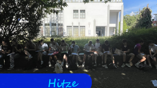 Vor Büschen auf Sitzbänken sitzen Jugendliche mit Büchern und Heften auf den Knien. Der Platz ist sehr schattig.