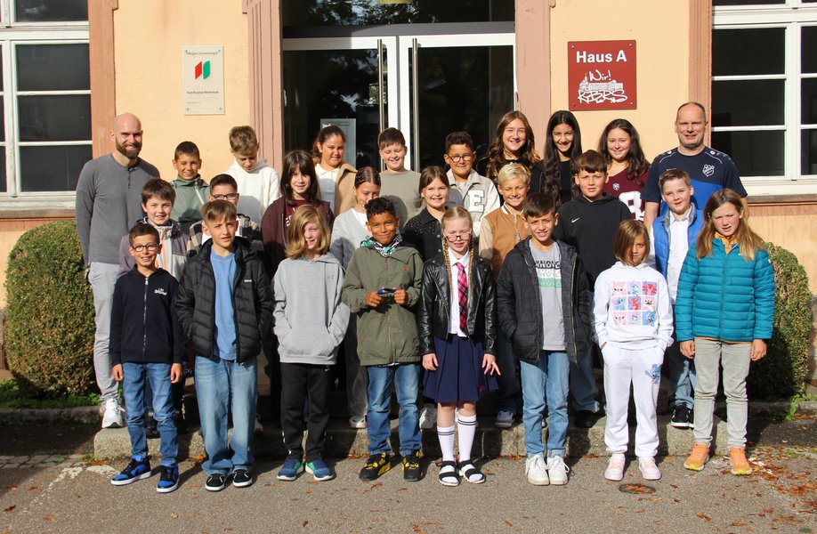 Vor dem Eingang der Schule hat sich eine fünfte Klasse mit den Klassenlehrkräften zum Gruppenfoto aufgestellt.