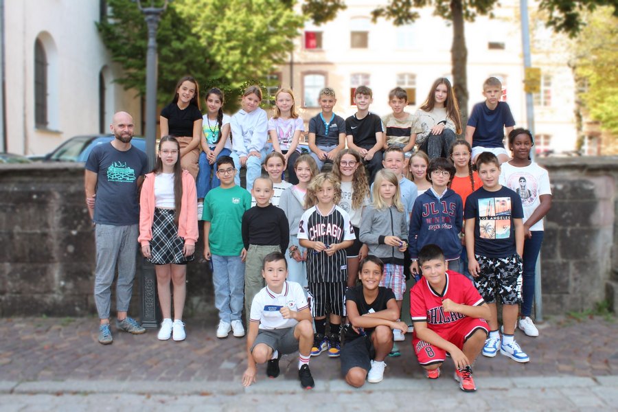 Schülerinnen und Schüler stehen für ein Klassenfoto zusammen. Die vordere Reihe kniet, dahinter steht eine Reihe die letzte Reihe sitzt auf einer Mauer. Im Hintergrund sieht man die Schule.