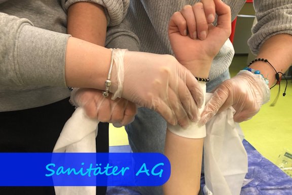 An einem nach oben gestreckten Arm sieht man zwei Hände in Einweghandschuhen einen Verband anlegen.
