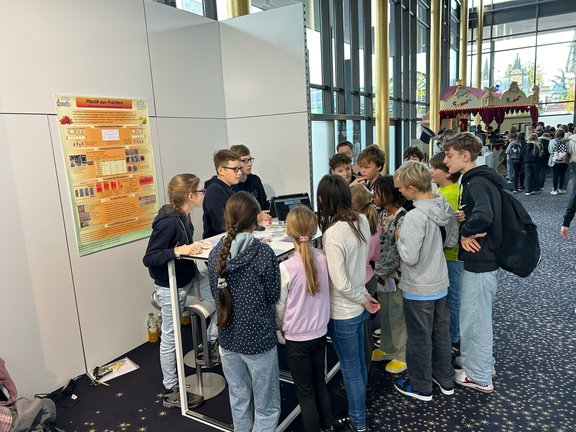 Hinter einem Auslagetisch stehen ein Mädchen und zwei Jungen, die den vor dem Tisch stehenden Jugendlichen Erklärungen abgeben. An der Wand im Hintergrund ist ein Plakat zur Erklärung zu sehen.