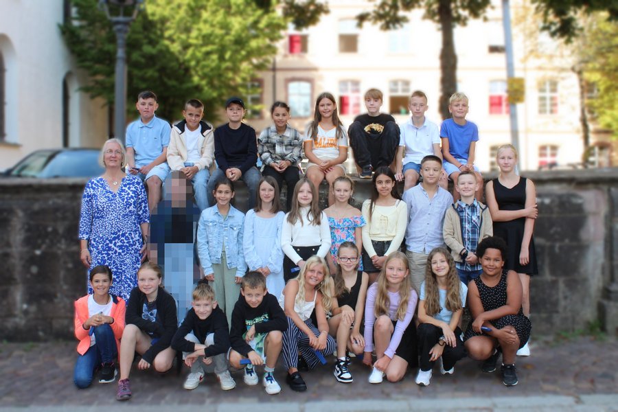 Schülerinnen und Schüler stehen für ein Klassenfoto zusammen. Die vordere Reihe kniet, dahinter steht eine Reihe die letzte Reihe sitzt auf einer Mauer. Im Hintergrund sieht man die Schule.