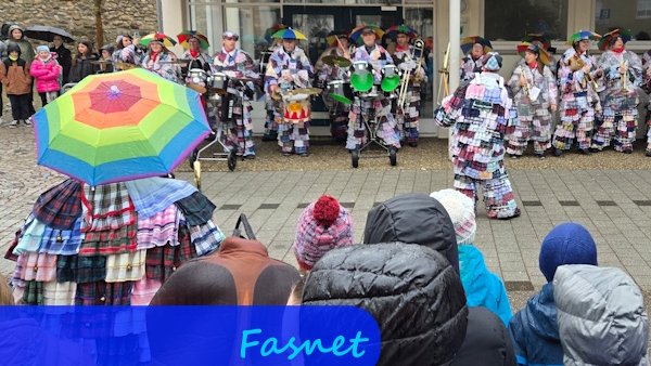 Unter einem Vordach stehen bunt verkleidete Menschen mit Blas- und Schlaginstrumenten und spielen Musik. Im Vordergrund sieht man die Rücken der zuschauenden Kinder und eine verkleidete Person mit buntem Regenschirm.