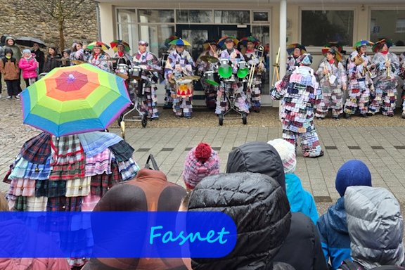 Unter einem Vordach stehen bunt verkleidete Menschen mit Blas- und Schlaginstrumenten und spielen Musik. Im Vordergrund sieht man die Rücken der zuschauenden Kinder und eine verkleidete Person mit buntem Regenschirm.