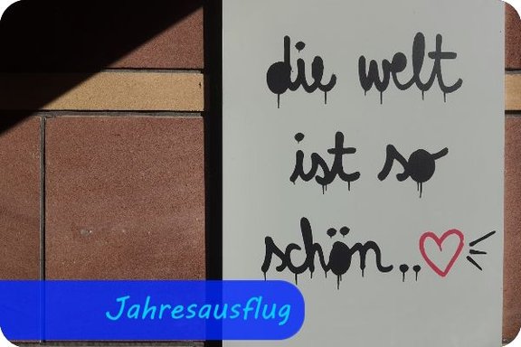 An einer Mauer hängt ein weißes Bild. Darauf stehen "Die Welt ist so schön..." und ein kleines, rotes Herz.