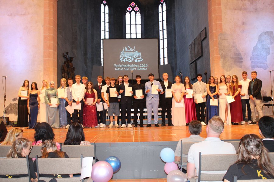 Auf der Bühne stehen weibliche und männliche Jugendliche mit Zeugnissen in der Hand zum Foto zusammen.
