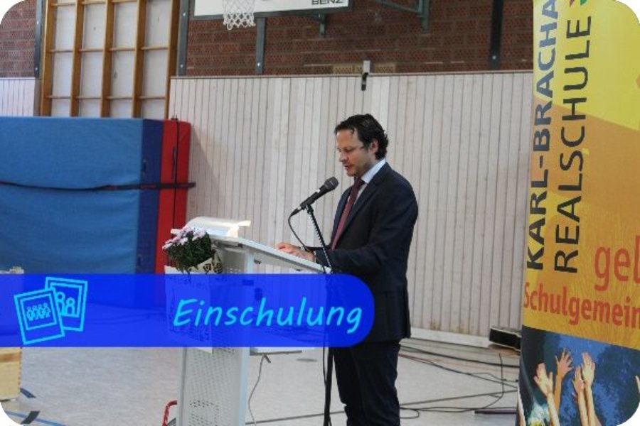 Eine männliche Person im Anzug steht an einem Rednerpult und spricht in eine Mikrofon. Am rechten Rand hängt ein Plakat mit der Aufschrift "Karl-Brachat-Realschule - gelebte Schulgemeinschaft". Überschrift des Bildes ist "Einschulung" mit einem Galeriesymbol.