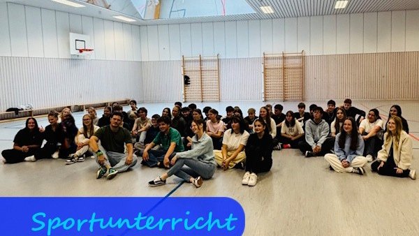 Auf dem Boden einer Turnhalle sitzen Mädchen und Jungen in legerer Sportkleidung.