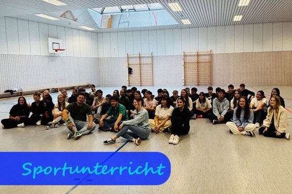 Auf dem Boden einer Turnhalle sitzen Mädchen und Jungen in legerer Sportkleidung.