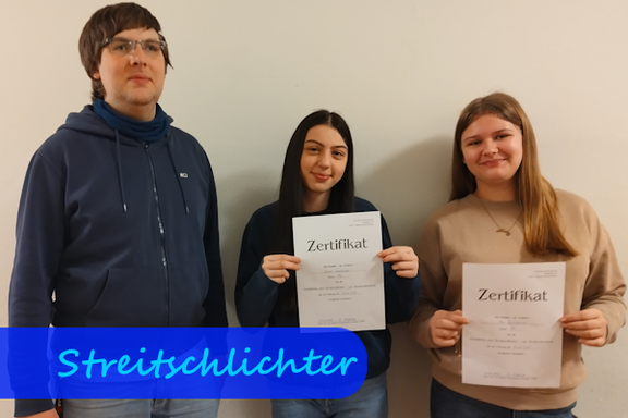 Von links nach rechts stehen ein Mann und zwei Mädchen. Die Mädchen halten ihr Zertifikat in die Kamera.