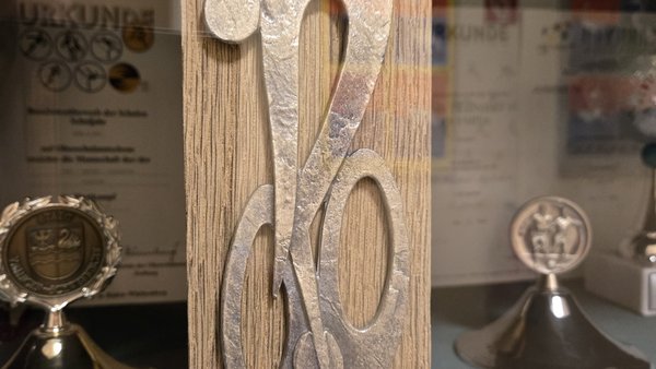 Auf einem hellen, senkrecht stehendem Holzbrett ist stilisiert ein Rennradfahrer aus Metall dargestellt. Am Fuß befindet sich ein Metallschild mit einer Gravur: Siegerklasse SCHULRADELN gestiftet vom Förderverein der KBRS 2024 Klasse 5a