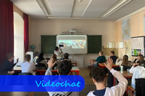 Aus den hinteren Reihen in einem Klassenzimmer sieht man vor sich Schülerinnen und Schüler, die sich melden, und vorne auf eine Videoleinwand schauen.