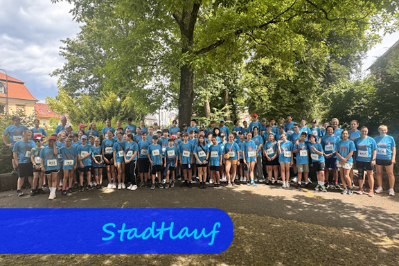 Vor einem großen Laubbaum stehen Kinder, Jugendliche und Erwachsen. Alle habe ein hellblaues Shirt an.