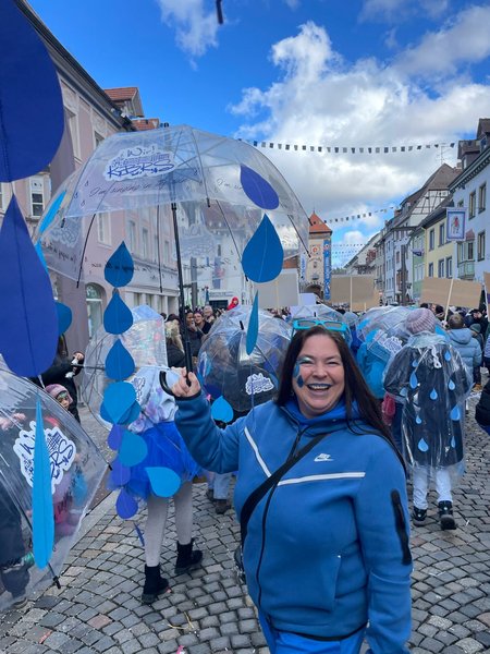 Während blau gekleidete Kinder mit transparenten Regenschirmen von der Kamera wegmarschieren, blickt eine blau gekleidete Frau umgedreht lächelnd zur Kamera und streckt den transparenten Regenschirm dem blauen Himmel entgegen.