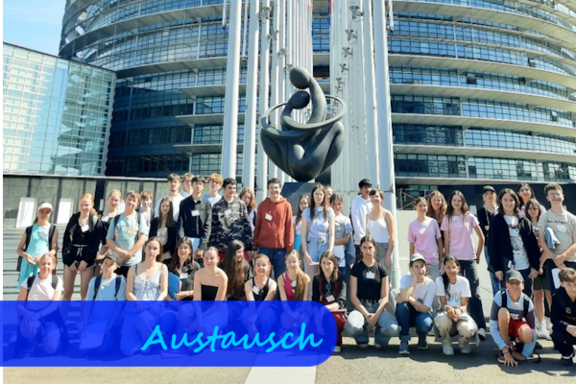 Eine Gruppe von Schülerinnen und Schülern schaut in die Kamera. Sie stehen vor einem runden Gebäude mit viel Glasfassade. Zwischen ihnen und dem Gebäude befinden sich eine graue Statue und viele Fahnenmasten.