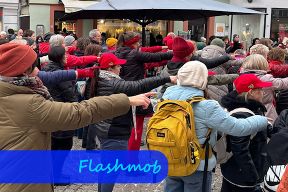 Viele Menschen, zum Teil mit roten Kopfbedeckungen, stehen nach rechts gewandt. Dabei strecken alle ihren Arm waagrecht nach vorn und zeigen auf etwas.