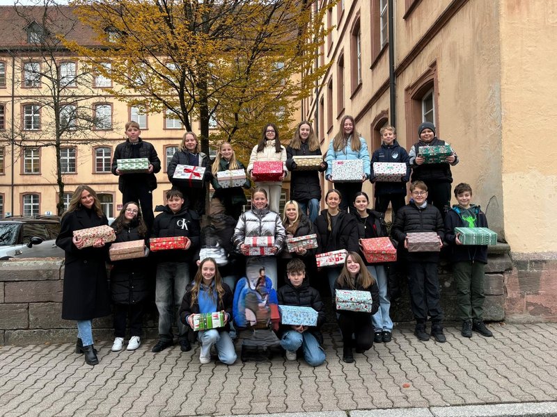 In drei Reihen stehen Schülerinnen und Schüler der Klassenstufe 7 vor und auf einer kleinen Mauer und schauen in die Kamera. In den Händen halten sie weihnachtlich verpackte Päckchen. Links daneben steht ihre Lehrerin, ebenfalls mit Päckchen in der Hand.