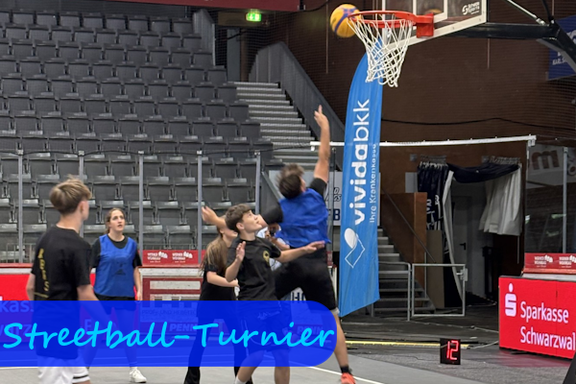 Sechs Jungen und Mädchen spielen Basketball. Ein Junge in blauem Trikot hat den Arm nach dem Wurf unter dem Korb noch erhoben, während der Basketball in Richtung des Korbes fliegt.